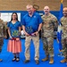 A group of five individuals pose for a formal photo in what appears to be a gymnasium or indoor facility. Four are wearing US Air Force OCP (Operational Camouflage Pattern) uniforms, while one civilian man in a blue polo shirt and one woman in a floral dress are also present. The civilian man is holding a decorative plaque featuring an Arizona state flag design, suggesting an award or recognition ceremony.