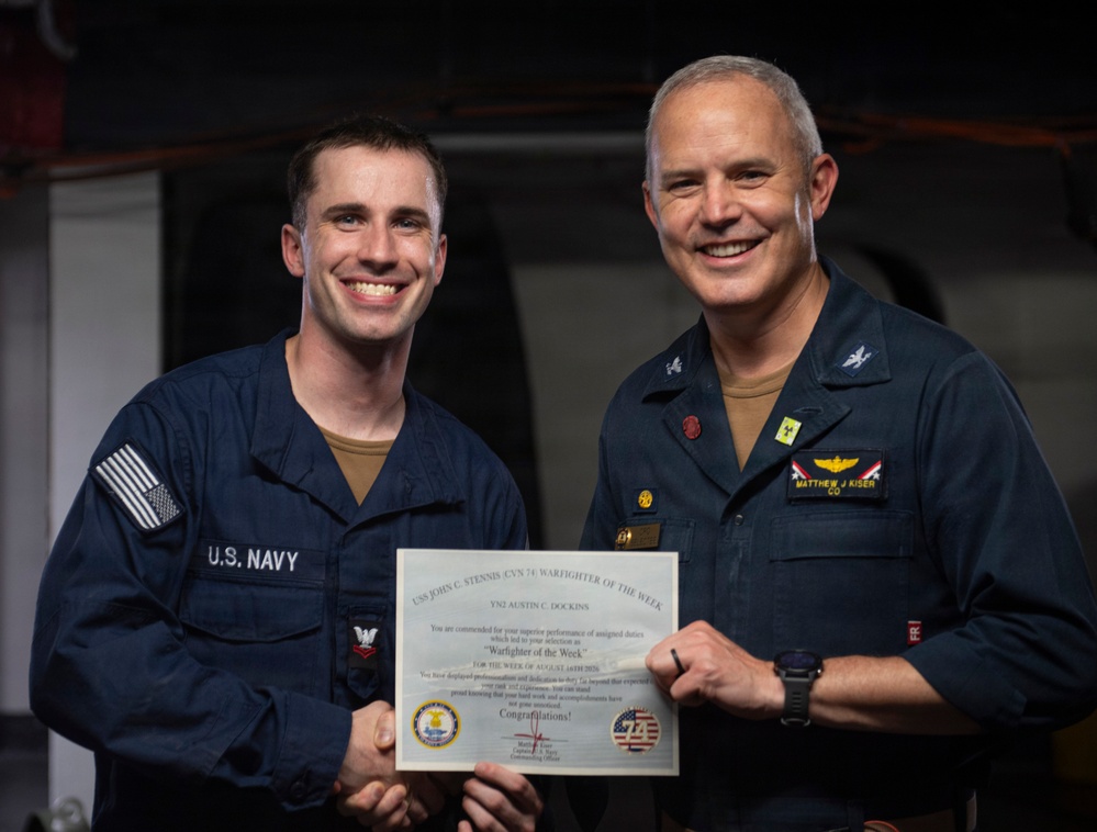 Two U.S. Navy personnel are posing together while holding a 'Warfighter of the Week' certificate aboard USS John C. Stennis (CVN 74). The officer on the right, identified by his name tag as Matthew J. Kiser with the rank insignia of a Captain (CO), is presenting the award to the sailor on the left, identified on the certificate as YN2 Austin C. Dockins. Both individuals are smiling and shaking hands for the commemorative photo.