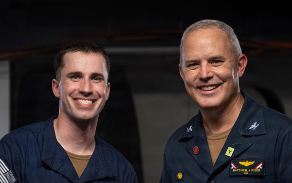Two U.S. Navy personnel are posing together while holding a 'Warfighter of the Week' certificate aboard USS John C. Stennis (CVN 74). The officer on the right, identified by his name tag as Matthew J. Kiser with the rank insignia of a Captain (CO), is presenting the award to the sailor on the left, identified on the certificate as YN2 Austin C. Dockins. Both individuals are smiling and shaking hands for the commemorative photo.
