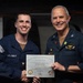 Two U.S. Navy personnel are posing together while holding a 'Warfighter of the Week' certificate aboard USS John C. Stennis (CVN 74). The officer on the right, identified by his name tag as Matthew J. Kiser with the rank insignia of a Captain (CO), is presenting the award to the sailor on the left, identified on the certificate as YN2 Austin C. Dockins. Both individuals are smiling and shaking hands for the commemorative photo.