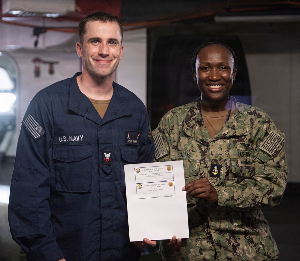 Two U.S. Navy personnel are posing together while holding a 'Warfighter of the Week' certificate. The sailor on the left, identified by his name tape as 'Austin Dockins,' is wearing Navy Working Uniform (NWU) Type III in blue, while the senior enlisted sailor on the right, identified as 'Hollis,' is wearing NWU Type III camouflage. The certificate appears to include a '1 Day Special Liberty' award.
