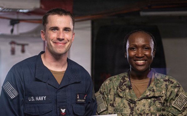 Two U.S. Navy personnel are posing together while holding a 'Warfighter of the Week' certificate. The sailor on the left, identified by his name tape as 'Austin Dockins,' is wearing Navy Working Uniform (NWU) Type III in blue, while the senior enlisted sailor on the right, identified as 'Hollis,' is wearing NWU Type III camouflage. The certificate appears to include a '1 Day Special Liberty' award.