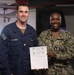 Two U.S. Navy personnel are posing together while holding a 'Warfighter of the Week' certificate. The sailor on the left, identified by his name tape as 'Austin Dockins,' is wearing Navy Working Uniform (NWU) Type III in blue, while the senior enlisted sailor on the right, identified as 'Hollis,' is wearing NWU Type III camouflage. The certificate appears to include a '1 Day Special Liberty' award.