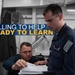A promotional military image showing two personnel in dark blue uniforms reviewing documents together aboard what appears to be a naval vessel. The standing individual is identified by a name tag as 'Austin Dockins' and wears a US flag patch on his sleeve. The image includes overlaid text reading 'WILLING TO HELP / READY TO LEARN,' suggesting a recruitment or training context.