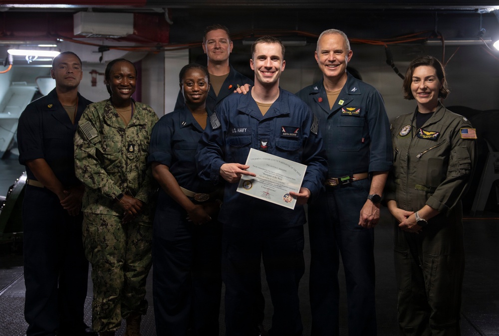 A group of seven U.S. Navy personnel pose for a formal photograph, with a central figure holding a certificate or award document. The setting appears to be aboard a naval vessel, likely in a hangar or below-deck space. The occasion appears to be a recognition or award ceremony.