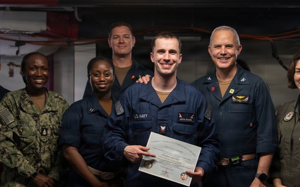 A group of seven U.S. Navy personnel pose for a formal photograph, with a central figure holding a certificate or award document. The setting appears to be aboard a naval vessel, likely in a hangar or below-deck space. The occasion appears to be a recognition or award ceremony.