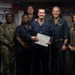 A group of seven U.S. Navy personnel pose for a formal photograph, with a central figure holding a certificate or award document. The setting appears to be aboard a naval vessel, likely in a hangar or below-deck space. The occasion appears to be a recognition or award ceremony.