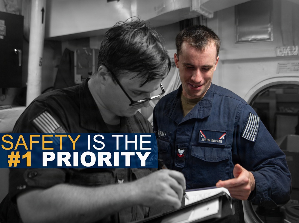 A U.S. Navy promotional safety poster featuring two sailors reviewing documents together in what appears to be a shipboard interior. The image uses selective color, with one sailor in full color wearing a blue Navy uniform with a name tag reading 'Austin Dockins,' while the background and other sailor are rendered in black and white. The overlay text reads 'SAFETY IS THE #1 PRIORITY.'