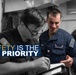 A U.S. Navy promotional safety poster featuring two sailors reviewing documents together in what appears to be a shipboard interior. The image uses selective color, with one sailor in full color wearing a blue Navy uniform with a name tag reading 'Austin Dockins,' while the background and other sailor are rendered in black and white. The overlay text reads 'SAFETY IS THE #1 PRIORITY.'