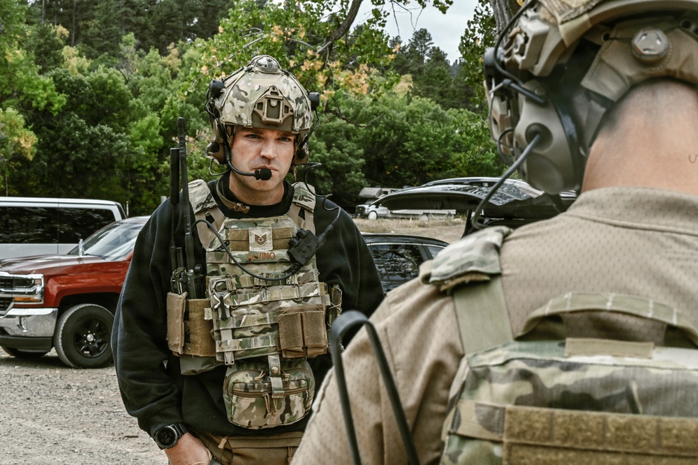 Two military personnel in full tactical gear are engaged in a face-to-face conversation in an outdoor setting surrounded by trees and parked vehicles. The central figure wears a multicam plate carrier with visible rank insignia and a tactical helmet equipped with communications equipment. The setting appears to be a staging or briefing area, likely during a training exercise or operational deployment.