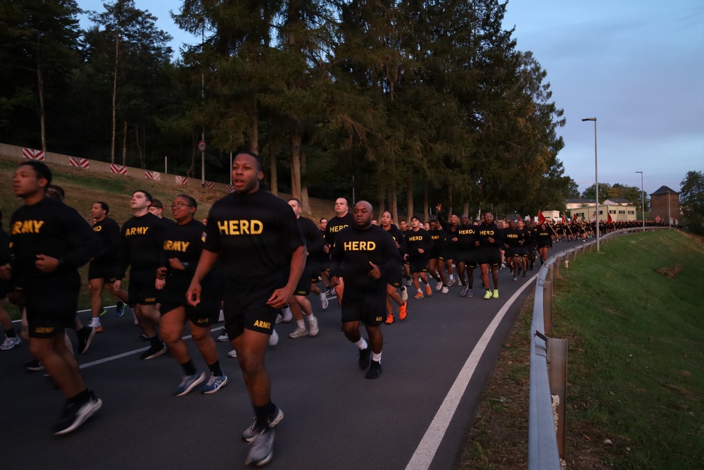 A large formation of U.S. Army personnel conducting a group physical training run along a paved road at dawn or dusk. Participants are wearing standard black Army PT (Physical Training) uniforms with gold lettering, with some shirts reading 'HERD' and others reading 'ARMY'. The formation stretches far into the background, suggesting a large-scale unit run or motivational run event.