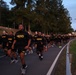 A large formation of U.S. Army personnel conducting a group physical training run along a paved road at dawn or dusk. Participants are wearing standard black Army PT (Physical Training) uniforms with gold lettering, with some shirts reading 'HERD' and others reading 'ARMY'. The formation stretches far into the background, suggesting a large-scale unit run or motivational run event.