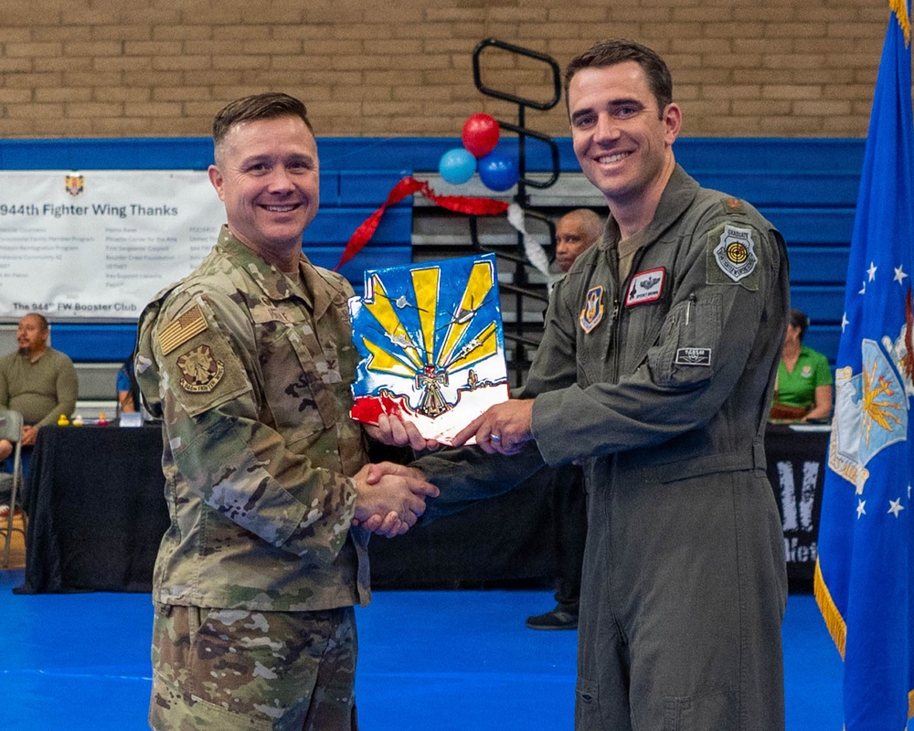 Two military personnel are posing together at a formal ceremony, jointly holding a colorful plaque or award featuring what appears to be the Arizona state flag design with sunburst rays in blue and yellow. The event appears to be associated with the 944th Fighter Wing, as indicated by signage visible in the background. The ceremony is taking place in what appears to be a gymnasium or indoor facility decorated with red, white, and blue balloons.