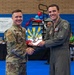 Two military personnel are posing together at a formal ceremony, jointly holding a colorful plaque or award featuring what appears to be the Arizona state flag design with sunburst rays in blue and yellow. The event appears to be associated with the 944th Fighter Wing, as indicated by signage visible in the background. The ceremony is taking place in what appears to be a gymnasium or indoor facility decorated with red, white, and blue balloons.