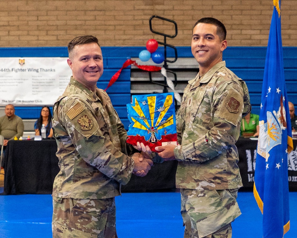 Two U.S. Air Force personnel in OCP (Operational Camouflage Pattern) uniforms are shaking hands while jointly holding a unit guidon or award plaque featuring a colorful emblem. The event appears to be a formal military ceremony taking place in a gymnasium, with a banner referencing the '944th Fighter Wing' visible in the background. An Air Force flag is displayed to the right of the subjects.