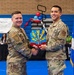 Two U.S. Air Force personnel in OCP (Operational Camouflage Pattern) uniforms are shaking hands while jointly holding a unit guidon or award plaque featuring a colorful emblem. The event appears to be a formal military ceremony taking place in a gymnasium, with a banner referencing the '944th Fighter Wing' visible in the background. An Air Force flag is displayed to the right of the subjects.