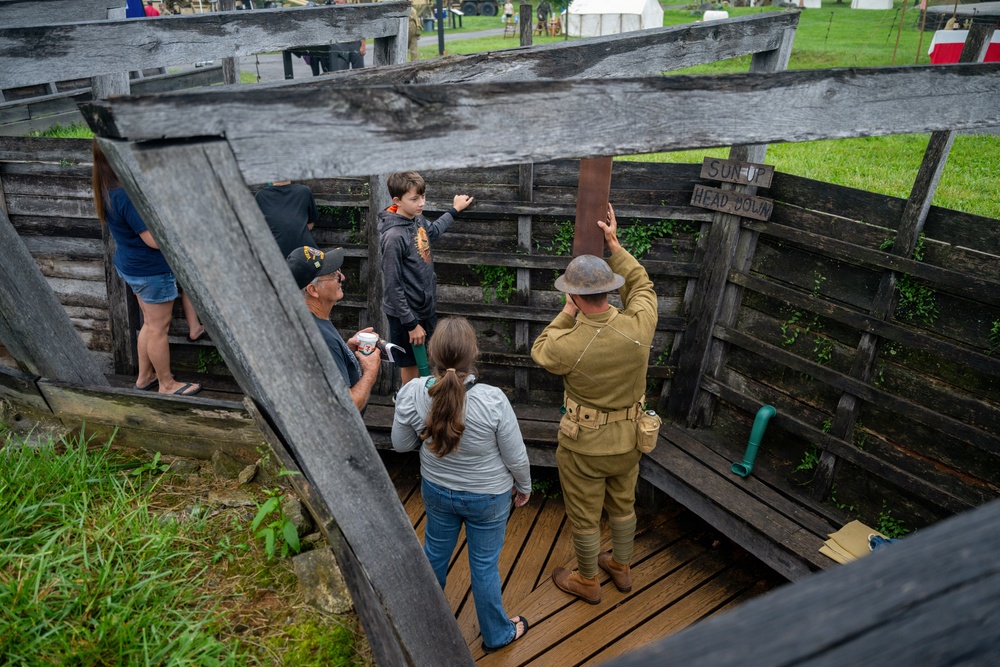 A WWI-era reenactor in period military uniform and helmet is guiding visitors through a reconstructed trench exhibit at what appears to be a living history event or museum. The reenactor is pointing to a sign reading 'SUN UP HEAD DOWN' on the wooden trench wall, demonstrating authentic trench warfare conditions. Visitors including adults and a child are gathered inside the wooden trench structure on a grassy outdoor site.