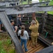 A WWI-era reenactor in period military uniform and helmet is guiding visitors through a reconstructed trench exhibit at what appears to be a living history event or museum. The reenactor is pointing to a sign reading 'SUN UP HEAD DOWN' on the wooden trench wall, demonstrating authentic trench warfare conditions. Visitors including adults and a child are gathered inside the wooden trench structure on a grassy outdoor site.