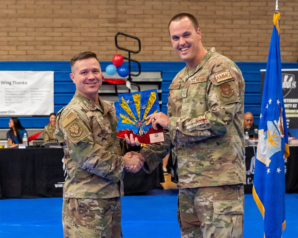 Two U.S. Air Force personnel in OCP (Operational Camouflage Pattern) uniforms are shaking hands while jointly holding an award or trophy during a formal recognition ceremony. The award appears to be a decorative plaque featuring the Arizona state flag design with aircraft motifs. The event is taking place in what appears to be a gymnasium or community center set up for a military awards ceremony.