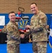 Two U.S. Air Force personnel in OCP (Operational Camouflage Pattern) uniforms are shaking hands while jointly holding an award or trophy during a formal recognition ceremony. The award appears to be a decorative plaque featuring the Arizona state flag design with aircraft motifs. The event is taking place in what appears to be a gymnasium or community center set up for a military awards ceremony.