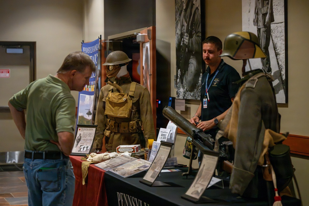 A military history exhibit, likely from the Pennsylvania Military Museum, displays World War I-era uniforms, equipment, and artifacts on a red-draped table. A staff member wearing a lanyard and badge engages with a visitor who is examining the displayed materials. Two mannequins dressed in period military uniforms and helmets flank the display table.