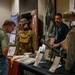 A military history exhibit, likely from the Pennsylvania Military Museum, displays World War I-era uniforms, equipment, and artifacts on a red-draped table. A staff member wearing a lanyard and badge engages with a visitor who is examining the displayed materials. Two mannequins dressed in period military uniforms and helmets flank the display table.
