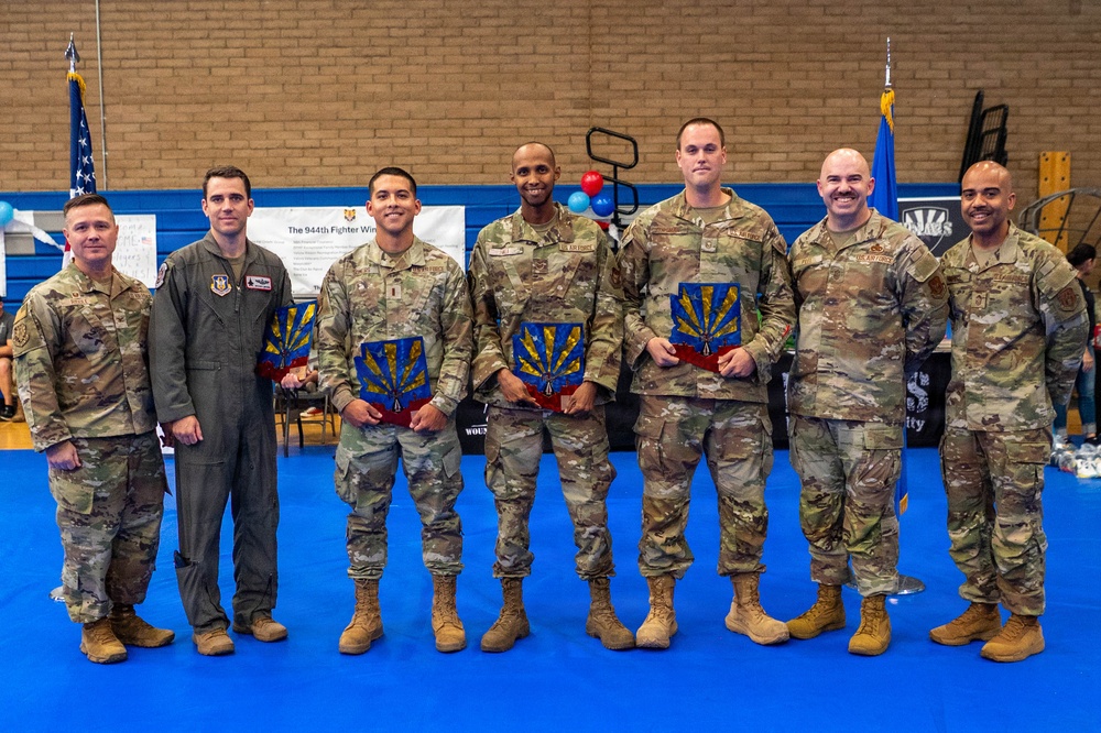 Seven military personnel pose for a group photo in what appears to be a gymnasium or indoor facility. Three individuals in the center are holding plaques or awards featuring what appears to be the Arizona state symbol in blue and gold. A banner in the background references the '944th Fighter Wing,' indicating this is a U.S. Air Force Reserve unit award ceremony.