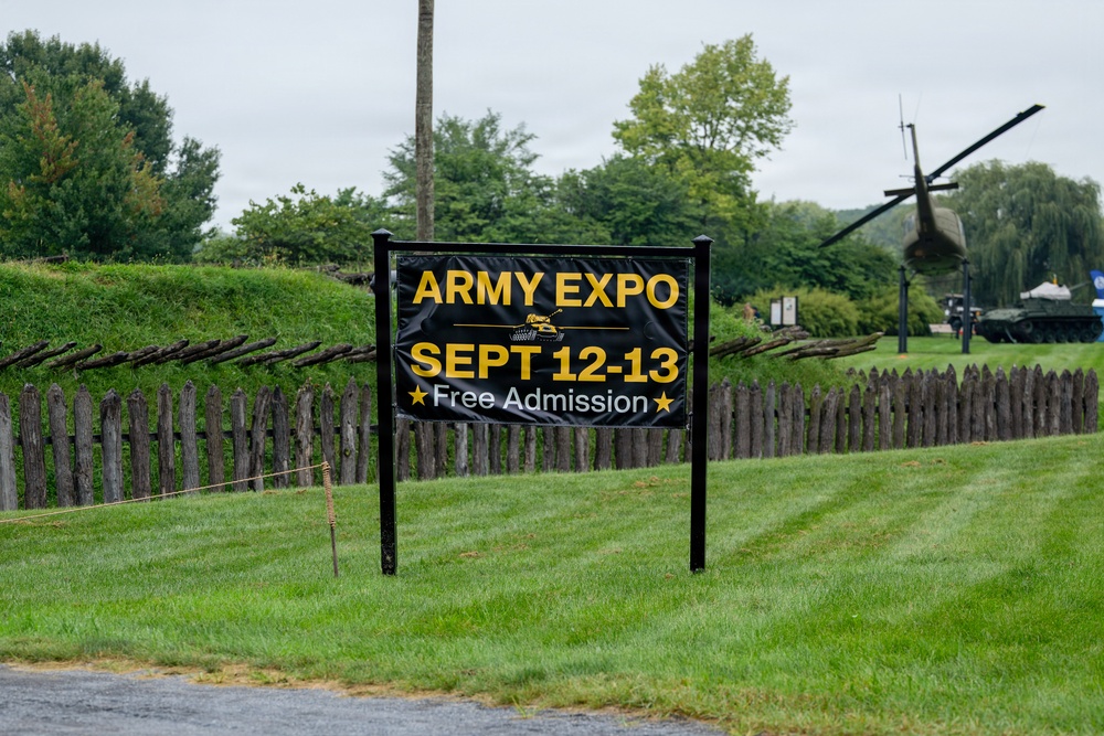 A black and yellow sign advertises an 'Army Expo' scheduled for September 12-13 with free admission. The sign is positioned along a well-maintained grassy area bordered by a rustic wooden picket fence. In the background, military equipment including what appears to be a helicopter and a tank are visible on display, suggesting this is a military museum or heritage site.