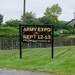 A black and yellow sign advertises an 'Army Expo' scheduled for September 12-13 with free admission. The sign is positioned along a well-maintained grassy area bordered by a rustic wooden picket fence. In the background, military equipment including what appears to be a helicopter and a tank are visible on display, suggesting this is a military museum or heritage site.