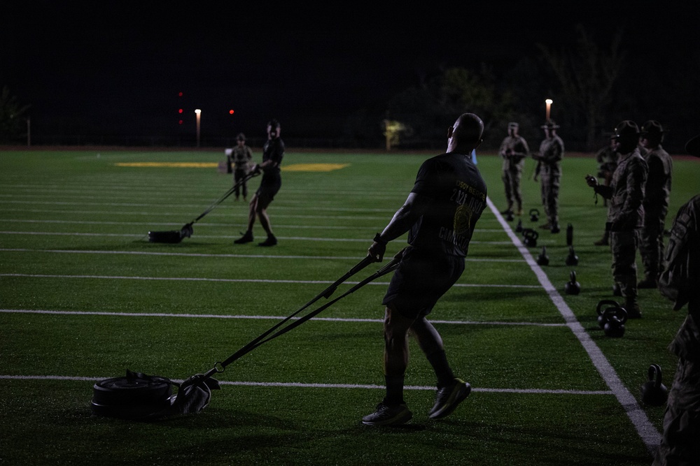 A nighttime outdoor physical training session is taking place on an artificial turf athletic field. Multiple individuals in military-style uniforms are engaged in various fitness exercises, with the primary subject in the foreground pulling a weighted sled using resistance straps. The scene is dimly lit by field lighting, creating a high-contrast, shadowed environment.