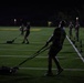 A nighttime outdoor physical training session is taking place on an artificial turf athletic field. Multiple individuals in military-style uniforms are engaged in various fitness exercises, with the primary subject in the foreground pulling a weighted sled using resistance straps. The scene is dimly lit by field lighting, creating a high-contrast, shadowed environment.