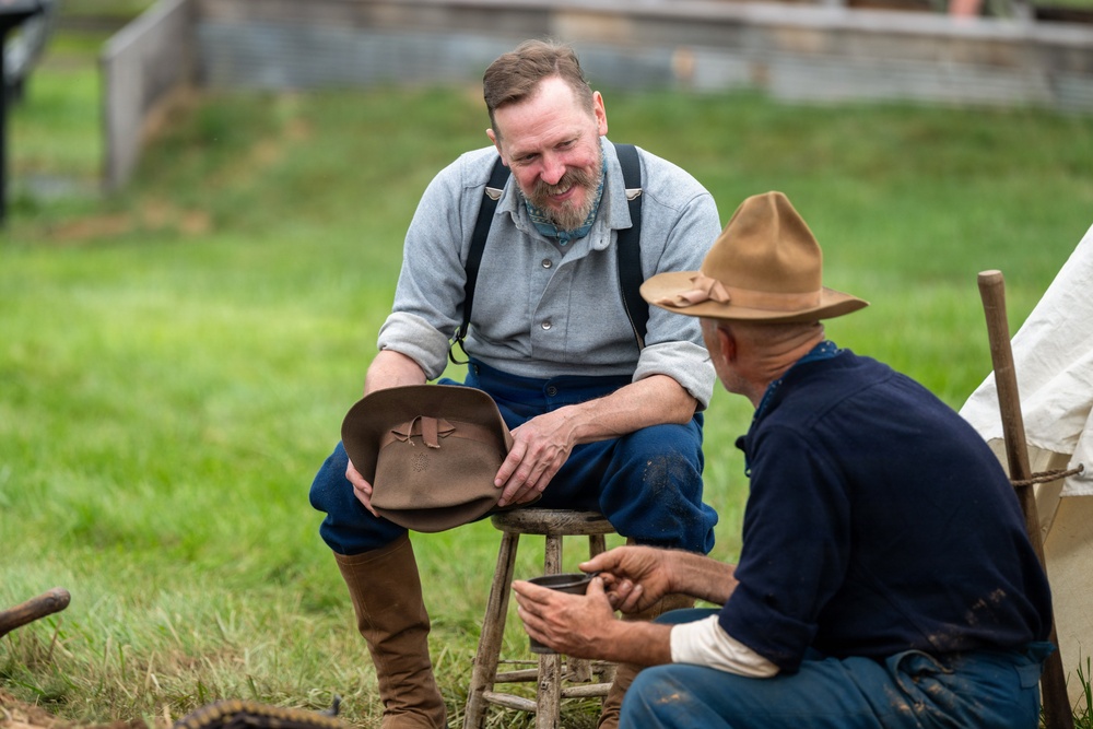Two men dressed in period-appropriate 19th-century clothing are engaged in a friendly conversation outdoors on a grassy field. The setting appears to be a historical reenactment, likely Civil War era, based on their attire including suspenders, work shirts, riding boots, and wide-brimmed felt hats. The man on the left is smiling and holding a hat while seated on a wooden stool.