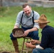 Two men dressed in period-appropriate 19th-century clothing are engaged in a friendly conversation outdoors on a grassy field. The setting appears to be a historical reenactment, likely Civil War era, based on their attire including suspenders, work shirts, riding boots, and wide-brimmed felt hats. The man on the left is smiling and holding a hat while seated on a wooden stool.