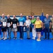 A group of approximately 13 individuals pose for a formal photo in what appears to be a military gymnasium or recreation facility. Several civilians are holding certificates or plaques, suggesting an awards or recognition ceremony. Military personnel in camouflage uniforms flank the group on both sides, and an American flag and Air Force flag are visible in the background.