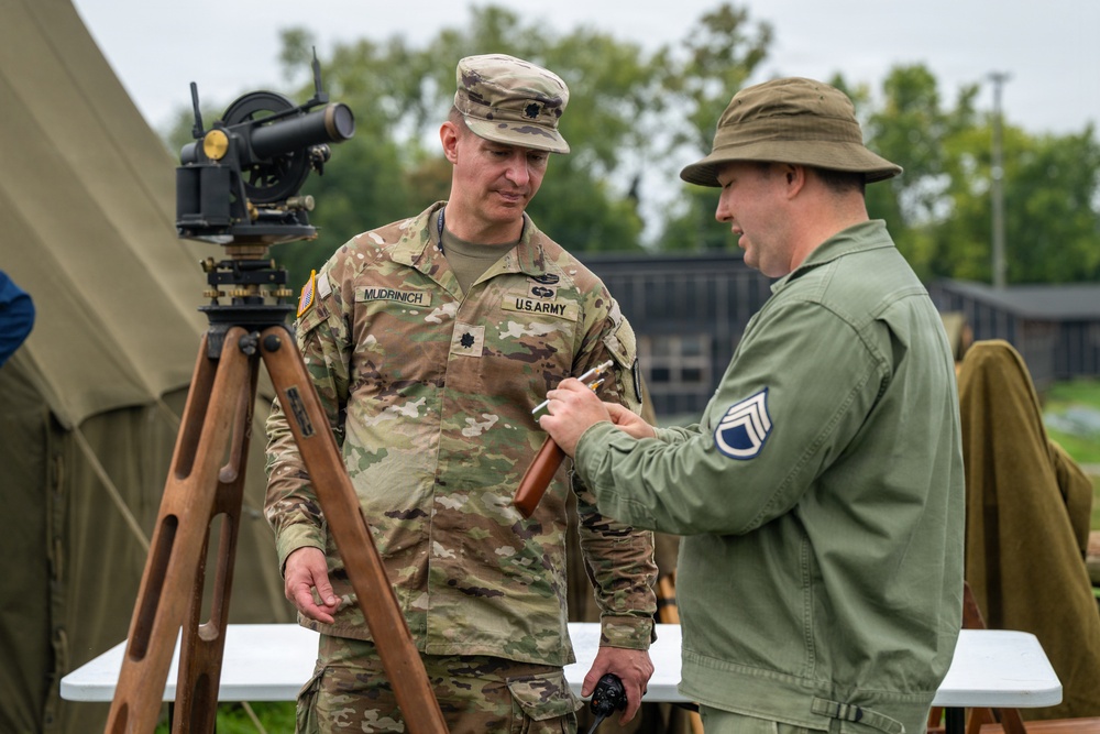 A U.S. Army Lieutenant Colonel named Mudrinich, wearing modern OCP (Operational Camouflage Pattern) uniform, is engaged in conversation with a person wearing vintage olive drab military attire bearing a Staff Sergeant chevron patch. They are standing near a vintage surveying instrument (theodolite) mounted on a wooden tripod, suggesting a historical demonstration or military heritage event. The setting includes military tents and outdoor tables consistent with a field display or commemorative event.
