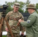 A U.S. Army Lieutenant Colonel named Mudrinich, wearing modern OCP (Operational Camouflage Pattern) uniform, is engaged in conversation with a person wearing vintage olive drab military attire bearing a Staff Sergeant chevron patch. They are standing near a vintage surveying instrument (theodolite) mounted on a wooden tripod, suggesting a historical demonstration or military heritage event. The setting includes military tents and outdoor tables consistent with a field display or commemorative event.