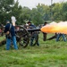A Civil War reenactment event is taking place on an open grassy field, with participants dressed in period-accurate Union and Confederate uniforms operating artillery cannons. One cannon is actively firing, producing a large orange flame and white smoke from the muzzle. A crowd of spectators is visible in the background watching the demonstration.