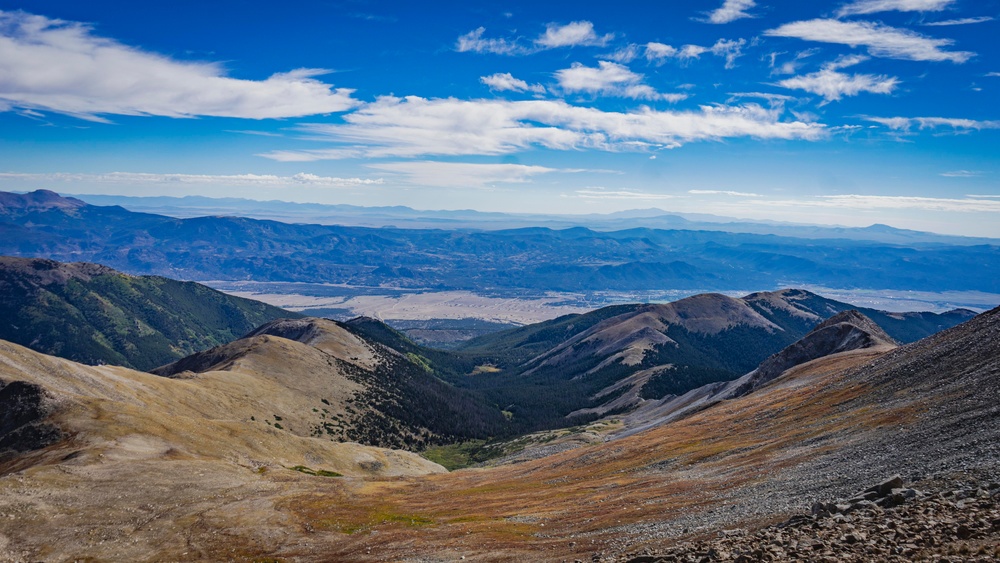 A high-altitude mountain panorama taken from a rocky summit or upper ridge, looking down into a broad valley with a distant urban or semi-arid basin visible in the middle ground. The foreground shows loose scree, talus, and sparse alpine vegetation transitioning to conifer-covered lower slopes. Multiple mountain ridges recede into a hazy blue horizon under a partly cloudy sky.