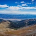 A high-altitude mountain panorama taken from a rocky summit or upper ridge, looking down into a broad valley with a distant urban or semi-arid basin visible in the middle ground. The foreground shows loose scree, talus, and sparse alpine vegetation transitioning to conifer-covered lower slopes. Multiple mountain ridges recede into a hazy blue horizon under a partly cloudy sky.