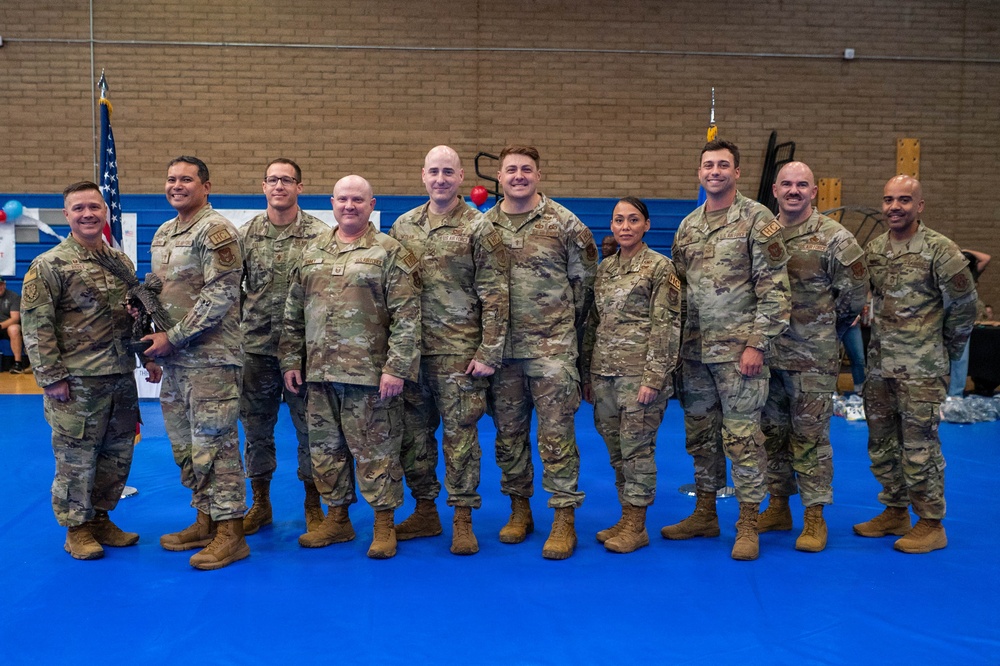 A group of approximately ten U.S. military personnel in OCP (Operational Camouflage Pattern) uniforms pose for a formal group photo inside what appears to be a gymnasium or indoor recreational facility. The group includes both male and female service members standing in a single row on a blue mat floor. The setting suggests a unit event, ceremony, or official gathering.