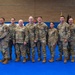A group of approximately ten U.S. military personnel in OCP (Operational Camouflage Pattern) uniforms pose for a formal group photo inside what appears to be a gymnasium or indoor recreational facility. The group includes both male and female service members standing in a single row on a blue mat floor. The setting suggests a unit event, ceremony, or official gathering.