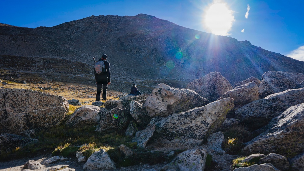 A hiker with a backpack stands on rocky terrain gazing up at a large, rugged mountain under a bright sun with visible lens flare. The landscape is high-altitude and alpine, characterized by scattered boulders, sparse grass patches, and a dark rocky peak. A second figure, possibly a person or dog, is seated among the rocks nearby.