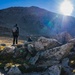 A hiker with a backpack stands on rocky terrain gazing up at a large, rugged mountain under a bright sun with visible lens flare. The landscape is high-altitude and alpine, characterized by scattered boulders, sparse grass patches, and a dark rocky peak. A second figure, possibly a person or dog, is seated among the rocks nearby.