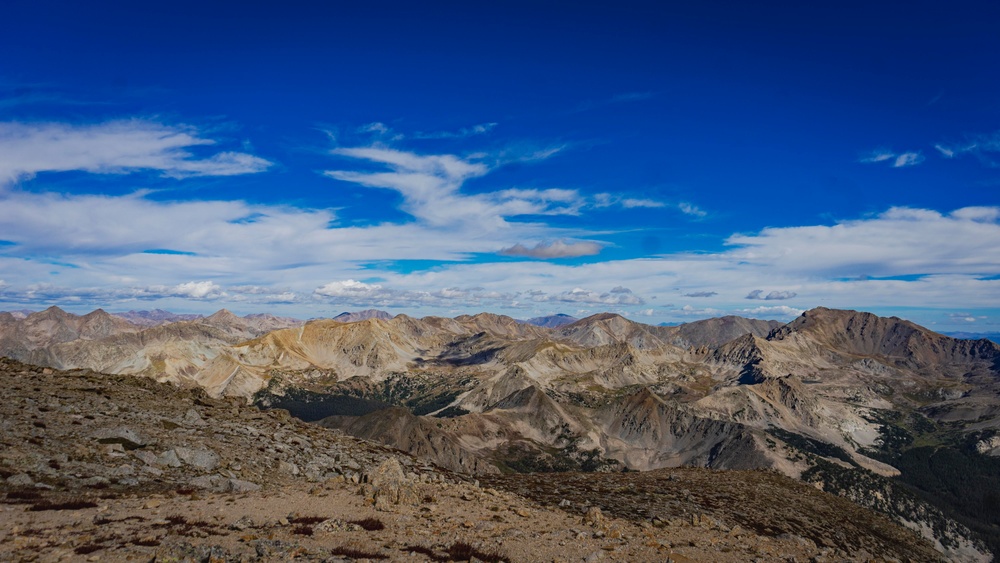 A panoramic high-altitude mountain landscape photographed from a summit or high ridge, showing an expansive range of rocky, barren peaks extending to the horizon. The terrain is predominantly brown and gray with minimal vegetation, suggesting an elevation above treeline. The deep blue sky with wispy cirrus and scattered cumulus clouds enhances the dramatic alpine scenery.