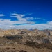 A panoramic high-altitude mountain landscape photographed from a summit or high ridge, showing an expansive range of rocky, barren peaks extending to the horizon. The terrain is predominantly brown and gray with minimal vegetation, suggesting an elevation above treeline. The deep blue sky with wispy cirrus and scattered cumulus clouds enhances the dramatic alpine scenery.