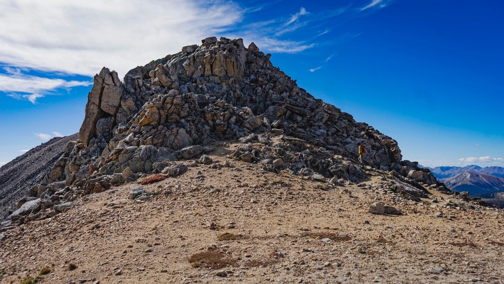 A high-altitude mountain summit scene showing a rocky peak under a vivid blue sky with wispy clouds. A lone hiker wearing a yellow jacket is visible near the base of the rocky summit formation, providing scale to the massive geological feature. The terrain is characterized by loose scree, sandy soil, and jagged exposed rock typical of alpine environments.