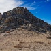 A high-altitude mountain summit scene showing a rocky peak under a vivid blue sky with wispy clouds. A lone hiker wearing a yellow jacket is visible near the base of the rocky summit formation, providing scale to the massive geological feature. The terrain is characterized by loose scree, sandy soil, and jagged exposed rock typical of alpine environments.