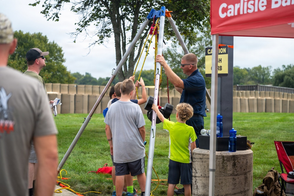 An adult in a navy blue shirt and sunglasses is assembling or demonstrating a tripod-style rescue/safety apparatus with several children assisting at an outdoor community event. The setup appears to be a confined space rescue tripod with rigging hardware at the top. A red 'Carlisle Ba[nk]' branded tent canopy is visible in the background, suggesting a sponsored public event.