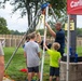 An adult in a navy blue shirt and sunglasses is assembling or demonstrating a tripod-style rescue/safety apparatus with several children assisting at an outdoor community event. The setup appears to be a confined space rescue tripod with rigging hardware at the top. A red 'Carlisle Ba[nk]' branded tent canopy is visible in the background, suggesting a sponsored public event.