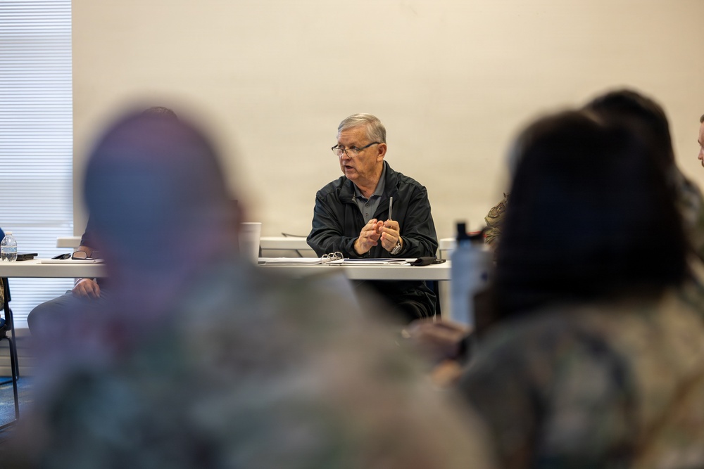 An older man with gray hair and glasses is seated at a table, appearing to address a group of people in what looks like a briefing or meeting room. Several attendees are visible in the foreground, some appearing to wear military camouflage uniforms. The speaker is holding a pen and gesturing with clasped hands, suggesting active discussion or presentation.
